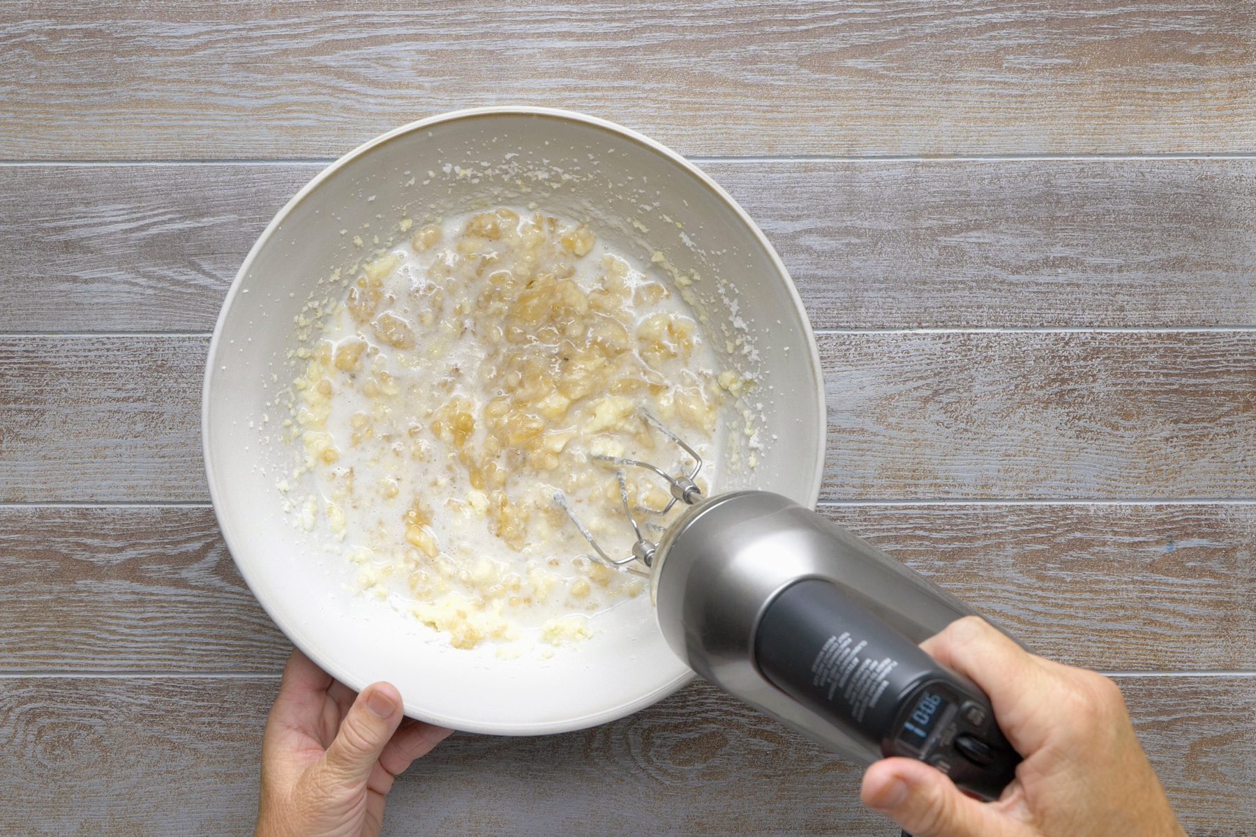 overhead shot; grey background; A white bowl contains a mixture of mashed bananas and a liquid, likely milk, An electric mixer is being used to beat the mixture together, creating a smooth and creamy consistency; The bananas are partially visible in the mixture