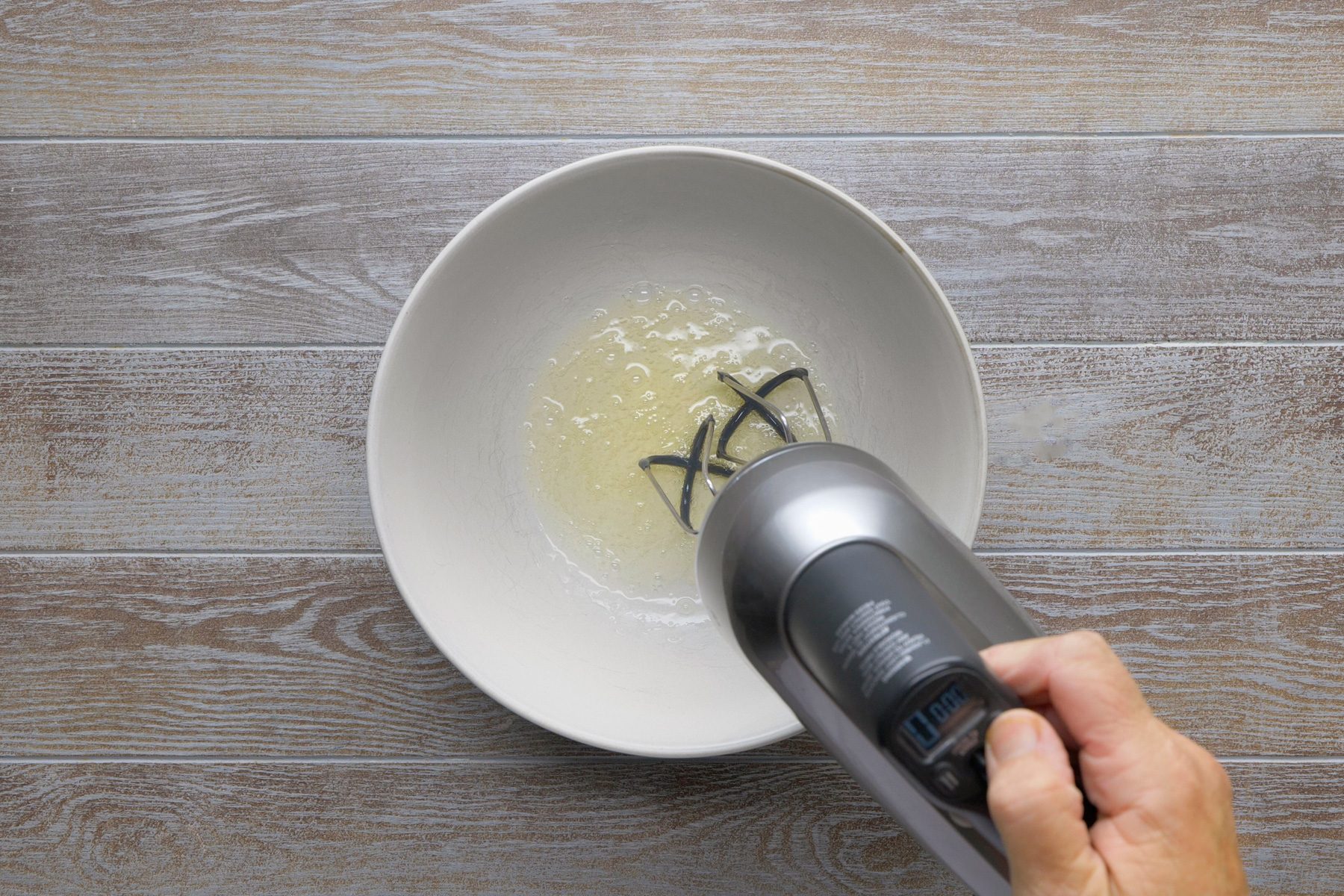 overhead shot; grey background; A hand is holding an electric mixer over a white bowl; The mixer is beating a liquid mixture, creating bubbles and foam on the surface; The liquid appears to be a light yellow color