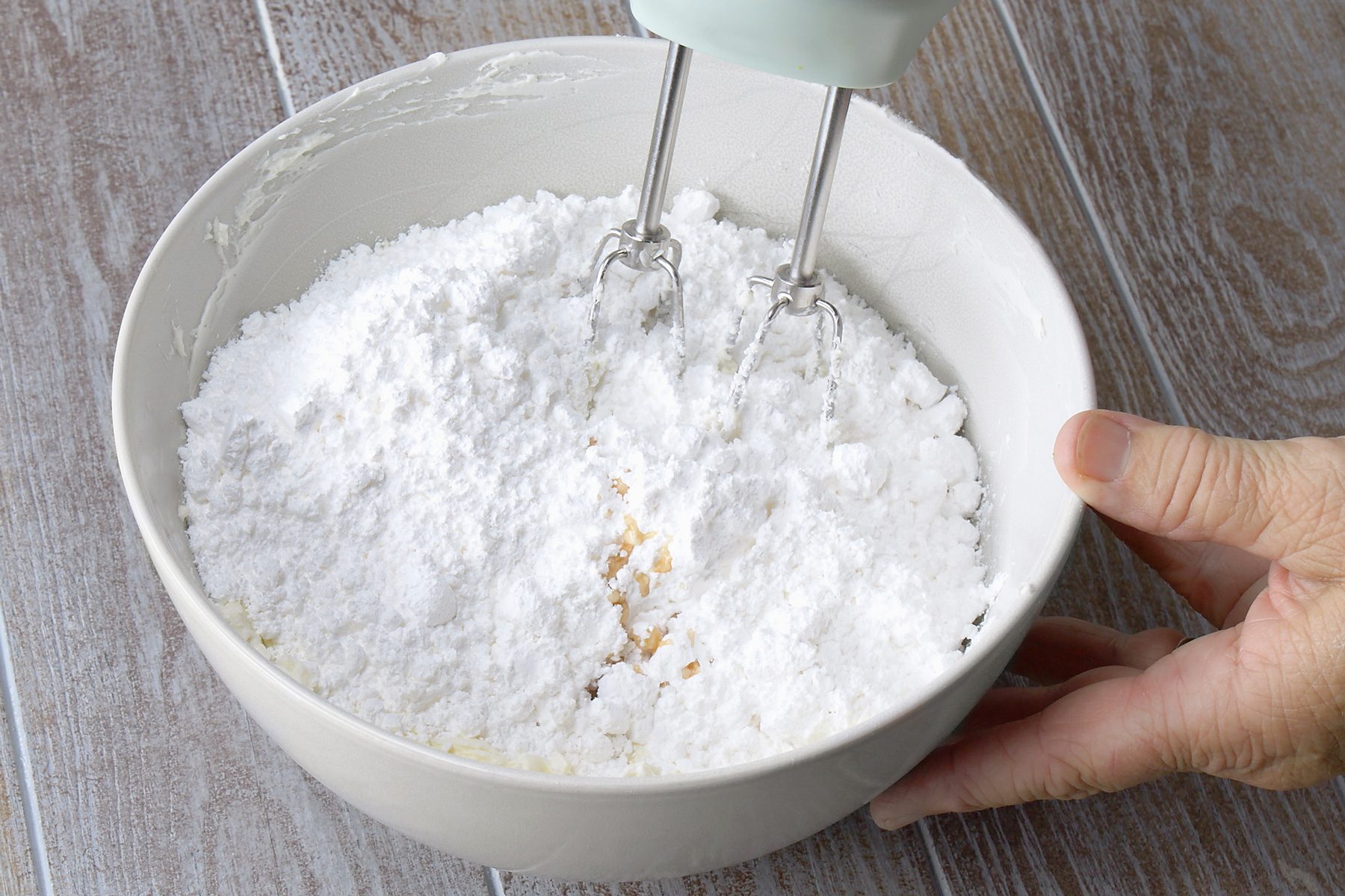 3/4th shot; grey background; A white bowl contains a mixture of powdered sugar, cream cheese, and a small amount of vanilla extract, An electric mixer is being used to beat the ingredients together, creating a creamy and fluffy frosting; The mixture is a pale yellow color, and the electric mixer is partially submerged in the bowl