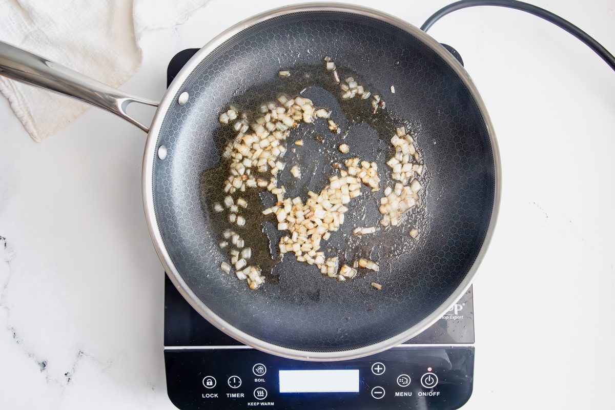 Sauted onions in a Hexclad pan with bacon grease for Taste of Home creamed cabbage on an induction cooktop on a marble surface.