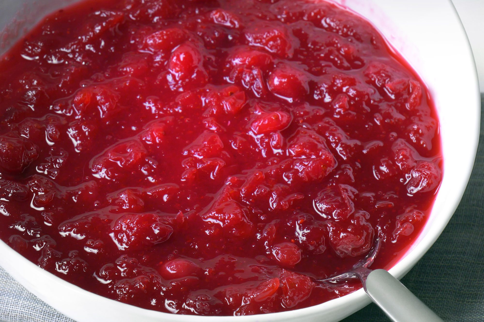 3/4th shot; A white bowl is filled with a vibrant red cranberry sauce, The sauce is thick and appears to have a smooth, slightly glossy texture, There are a few visible cranberry pieces within the sauce;