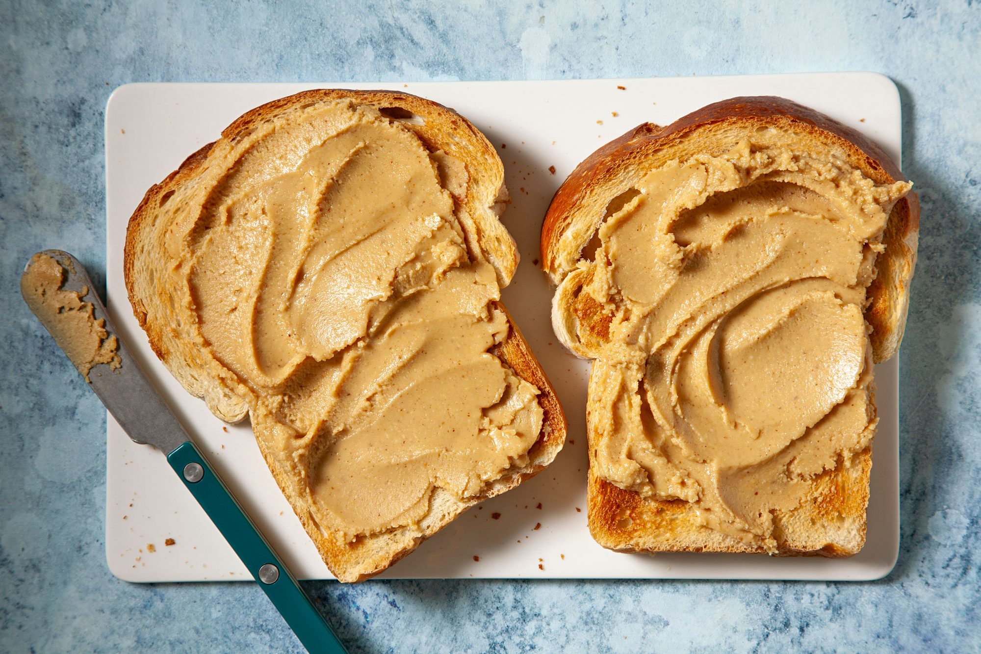 overhead shot; light blue textured background; Two slices of toasted bread are spread with a generous layer of creamy peanut butter; The bread slices are placed on a white cutting board, with a knife resting nearby;