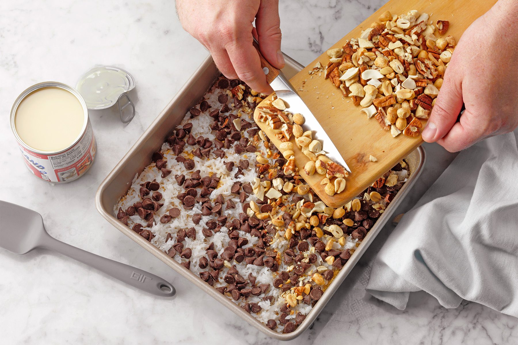 A person's hands are seen adding a mixture of nuts from a cutting board into a baking pan. The pan already contains a layer of chocolate chips and shredded coconut. A can of sweetened condensed milk and a spatula are on the marble countertop beside the pan.