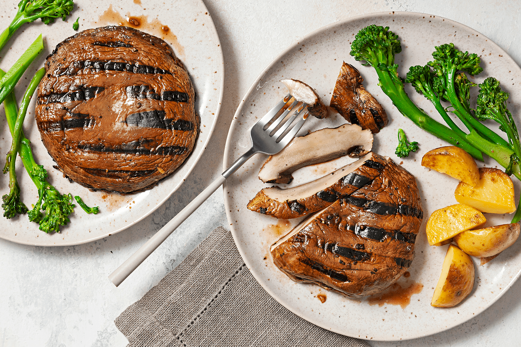 Two plates with grilled portobello mushrooms, each served with broccolini and roasted potato wedges. One plate shows a whole mushroom cap, while the other has slices of the mushroom with a fork. The plates are on a light surface with a grey napkin.