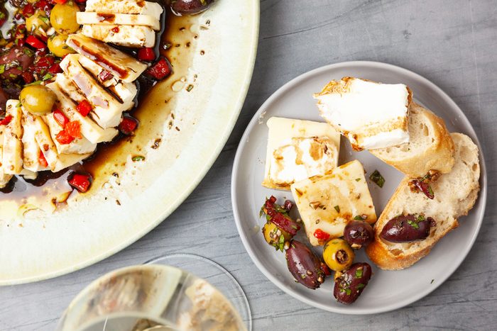 Close up of single servings of Taste of Home Olives & Cheese and full yield peakin in on a serving plate, sliced baguette on a cutting board, white wine