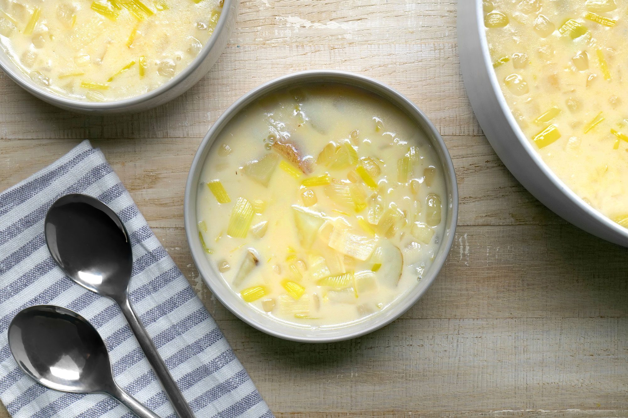 overhead shot; wooden background; A bowl of creamy Potato Leek Soup is the centerpiece of the image, The soup is a pale yellow color, with visible chunks of potato and leek; Three bowls of soup are arranged on a wooden table, along with two spoons and a blue and white striped napkin;