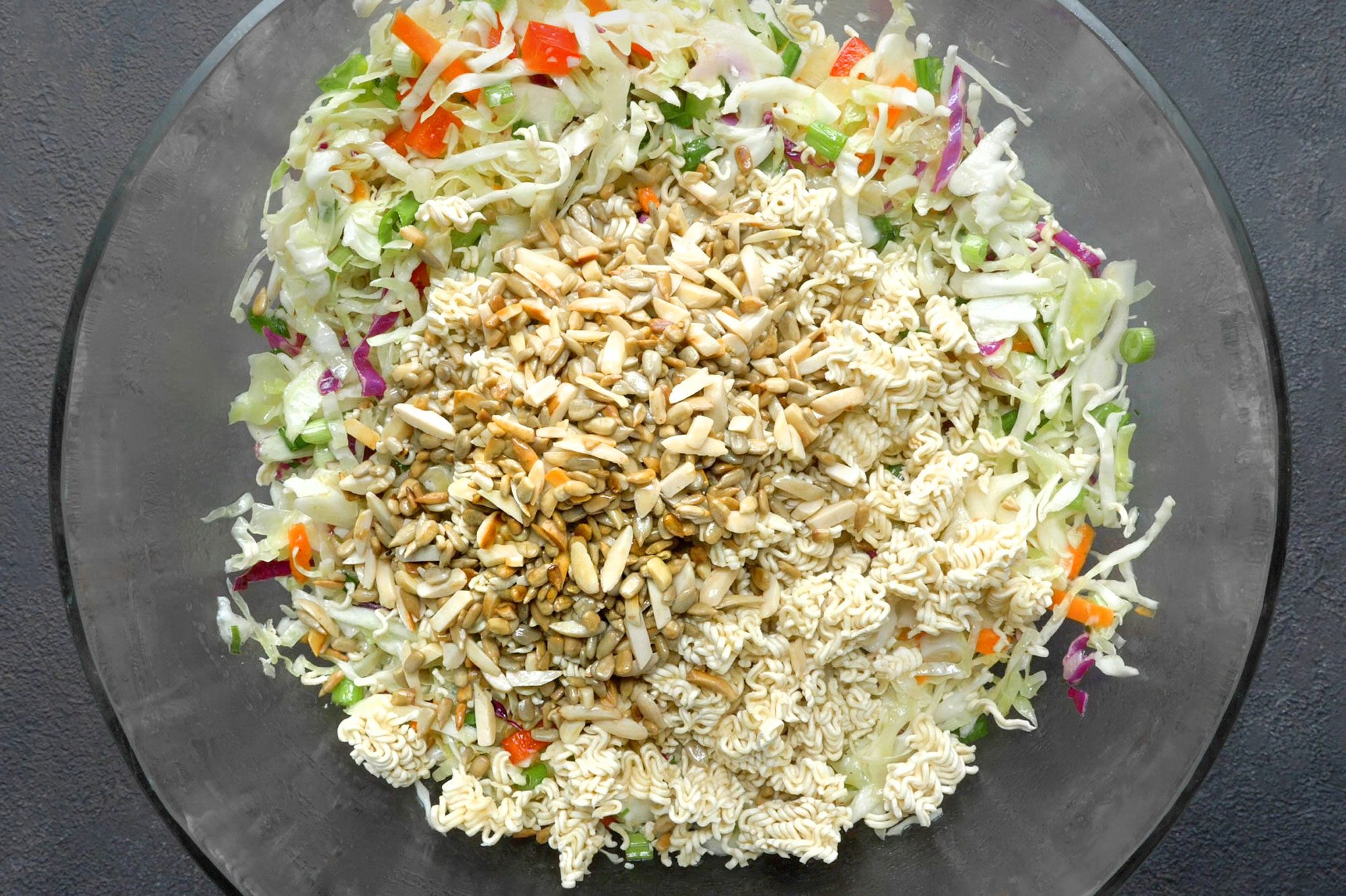 overhead shot; dark grey textured background; added noodles, almonds and sunflower kernels to the mixture in a large bowl;