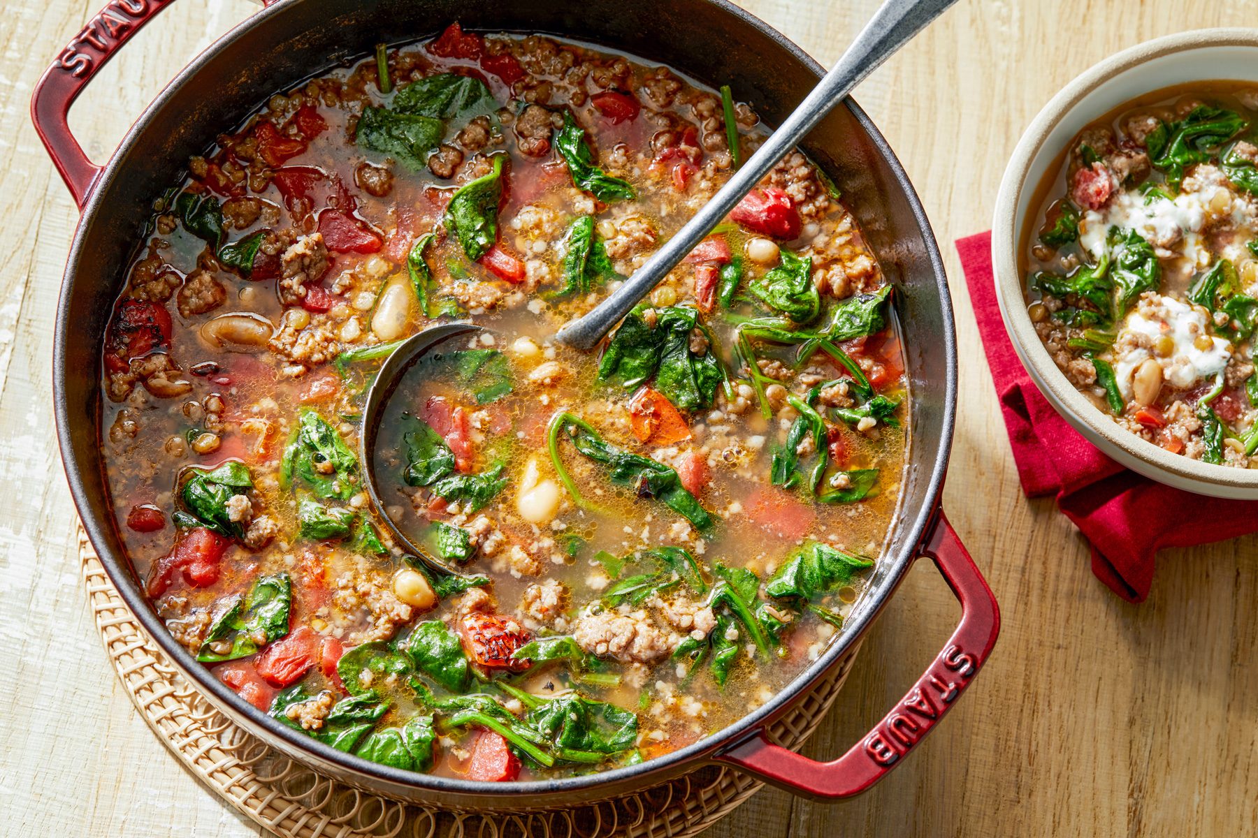 wooden background; A large, red Staub Dutch oven filled with Spinach and Sausage Lentil soup is the centerpiece of the image, A spoon rests in the soup, A smaller bowl filled with the soup and a dollop of crumbled feta cheese is visible in the background