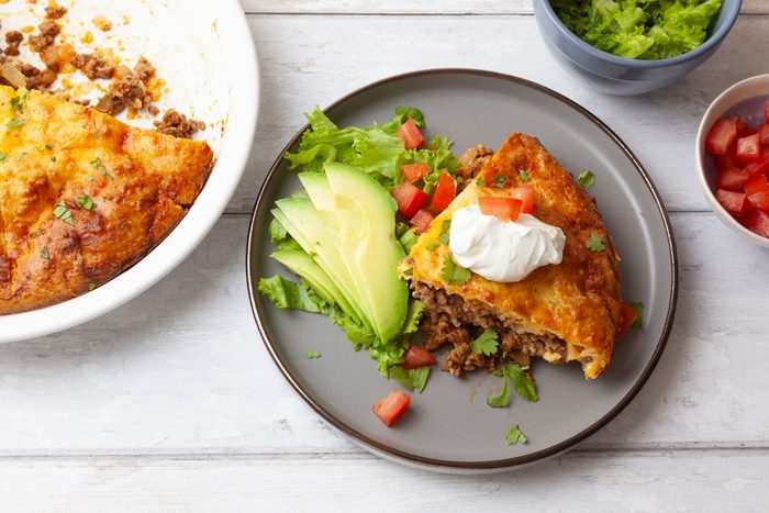Single serving of Taste of Home Taco Pie on a white wood surface with bowls of lettuce and tomatoes, full yield peaking in