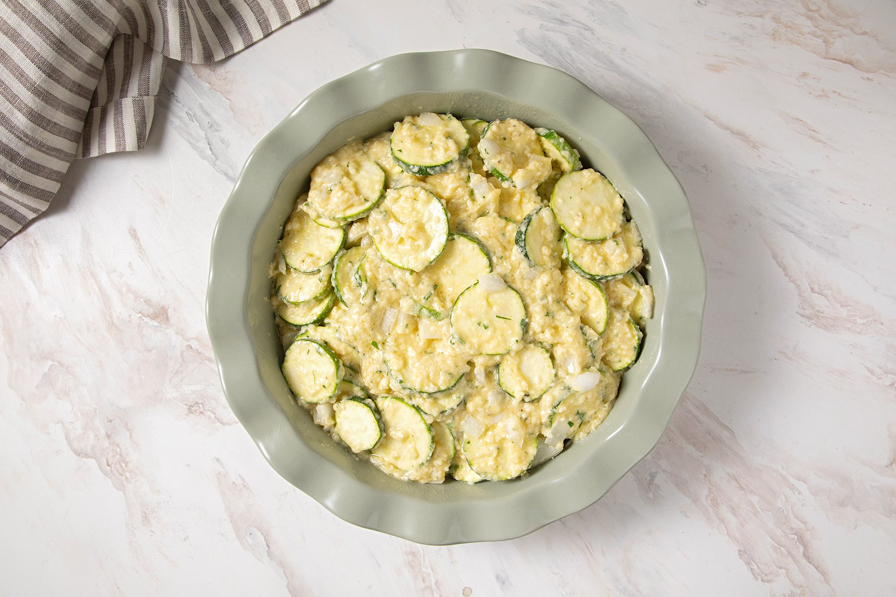 A top-down view of a round pie dish filled with a casserole featuring sliced zucchini and a creamy, golden mixture. Part of a striped cloth is seen in the top left corner on a marble countertop.