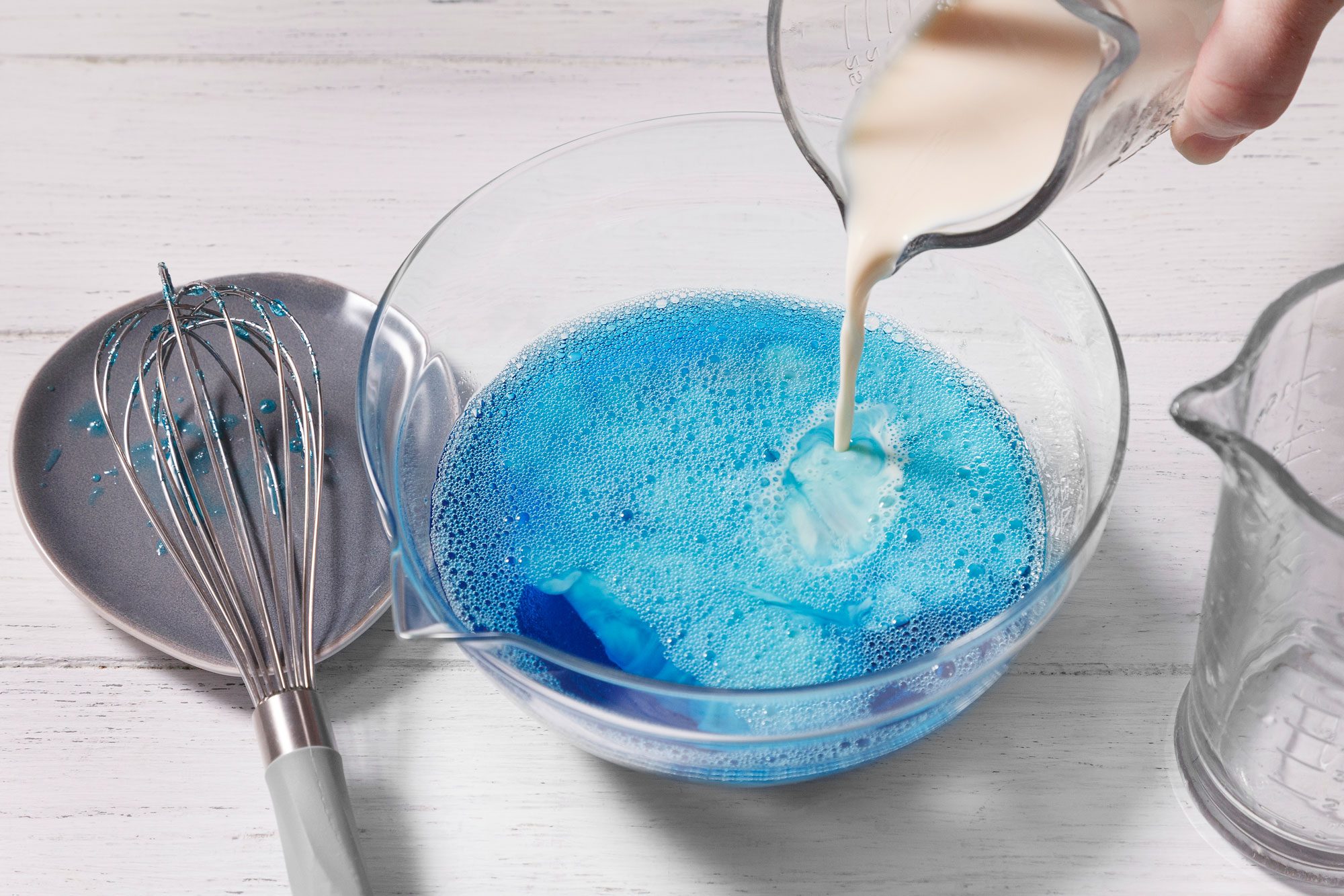 A glass measuring cup is pouring milk into a bowl of blue liquid