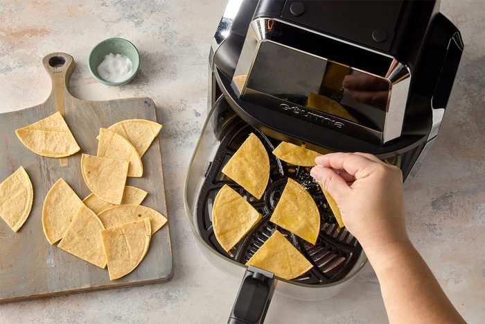 overhead shot of an air fryer with a basket full of tortilla chips, A person is sprinkling the chips with cooking salt, There is a wooden cutting board to the left with more tortilla chips;