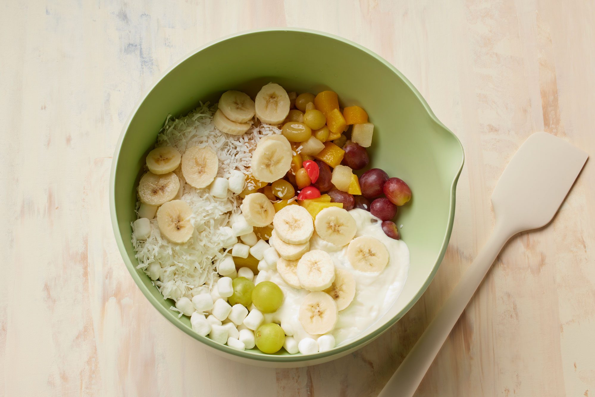 overhead shot of a large bowl placed over wooden background, combined all ingredients for Ambrosia Fruit Salad, a spatula is placed aside of a bowl;