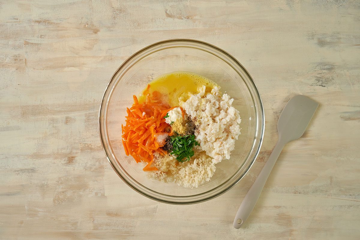 Overhead view of crabmeat, bread crumbs, shredded carrot, egg, melted butter, parsley, mayonnaise, Worcestershire sauce, mustard, salt, and pepper being gently folded together in a large bowl for the Taste of Home Baked Crab Cakes recipe.
