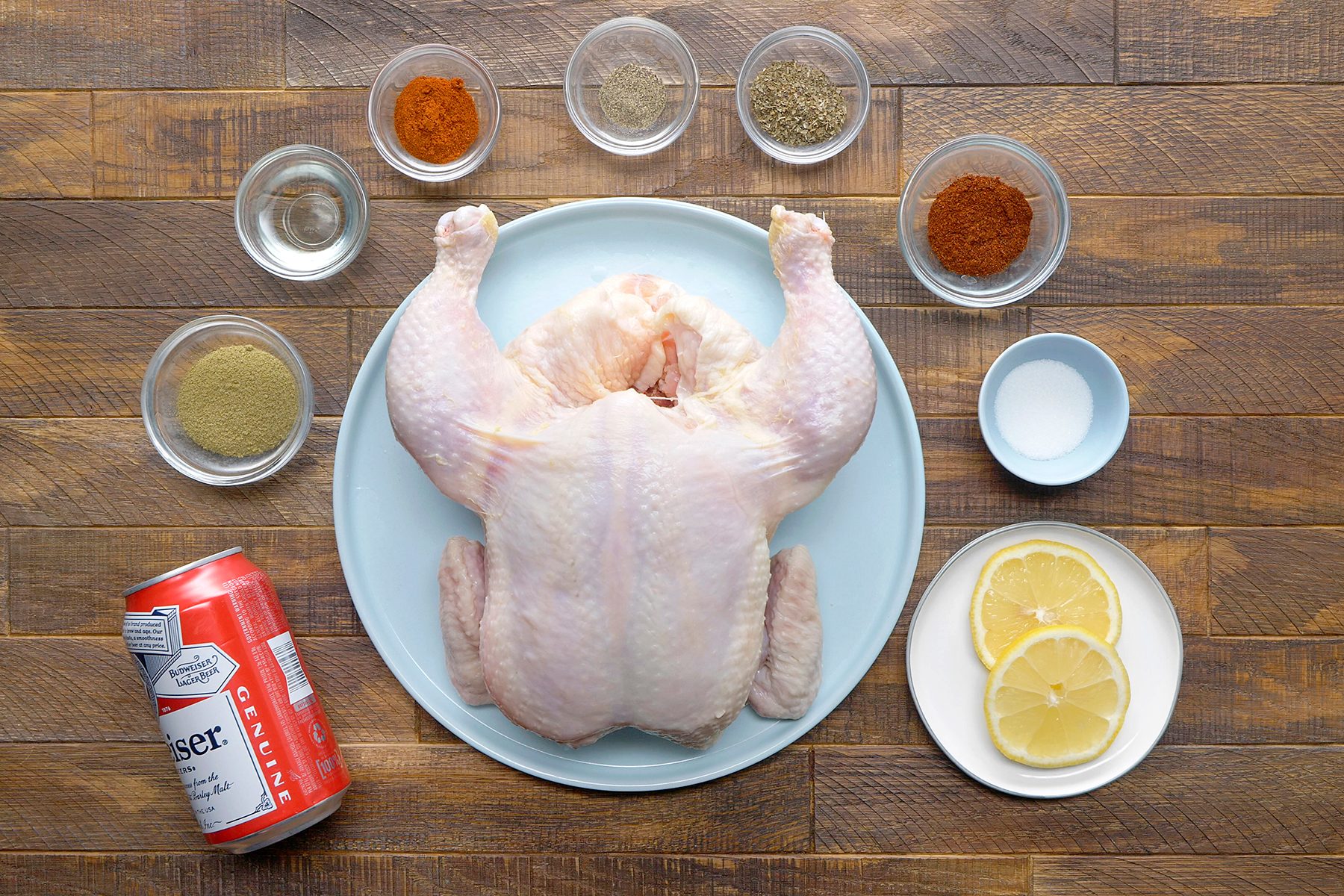 A raw whole chicken on a blue plate is surrounded by small bowls of various spices, two lemon slices on a white plate, a can of Budweiser beer, and a small bowl of salt, all placed on a wooden surface.
