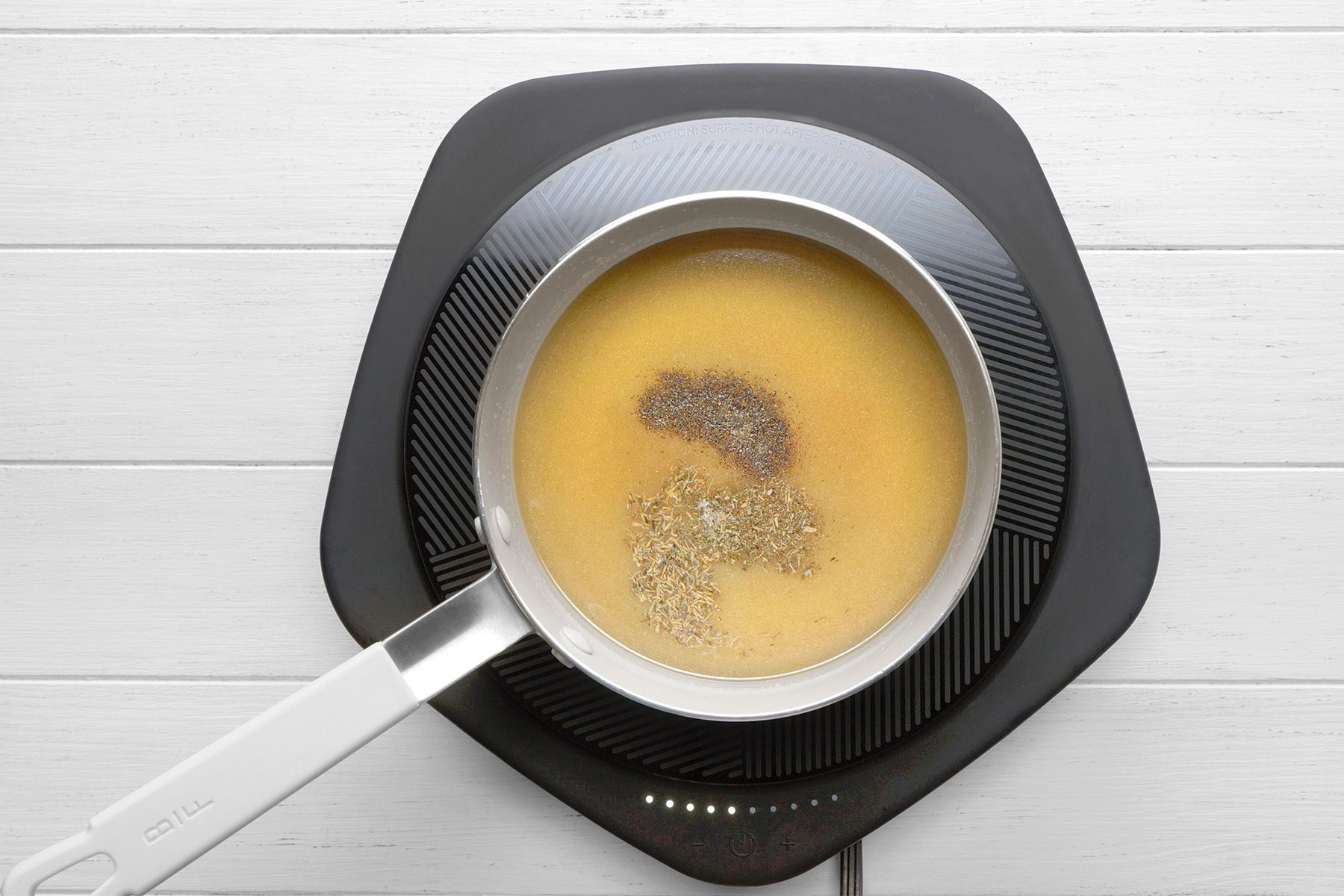 overhead shot of a saucepan on an electric induction cooktop, The saucepan contains a liquid that is a pale yellow color, A pinch of spices has been added to the liquid, creating a small dark spot in the center, The liquid is bubbling slightly, indicating that it is heating up;