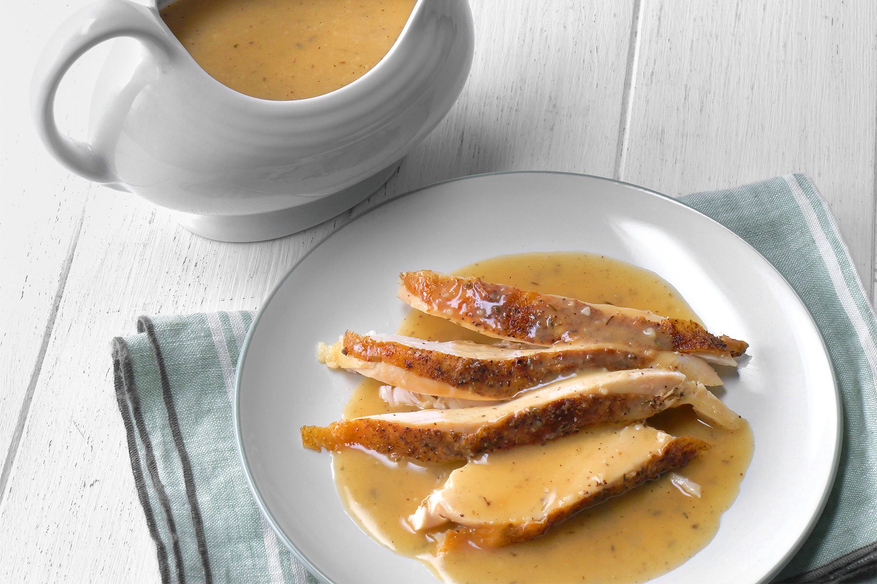 3/4th shot of a delicious dish of chicken gravy, The chicken is sliced and arranged neatly on a white plate, showcasing a golden-brown seared crust, Beside the plate, there’s a gravy boat filled with creamy, herb-speckled gravy, The dish is presented on a white wooden table with a folded green-striped napkin under the plate;