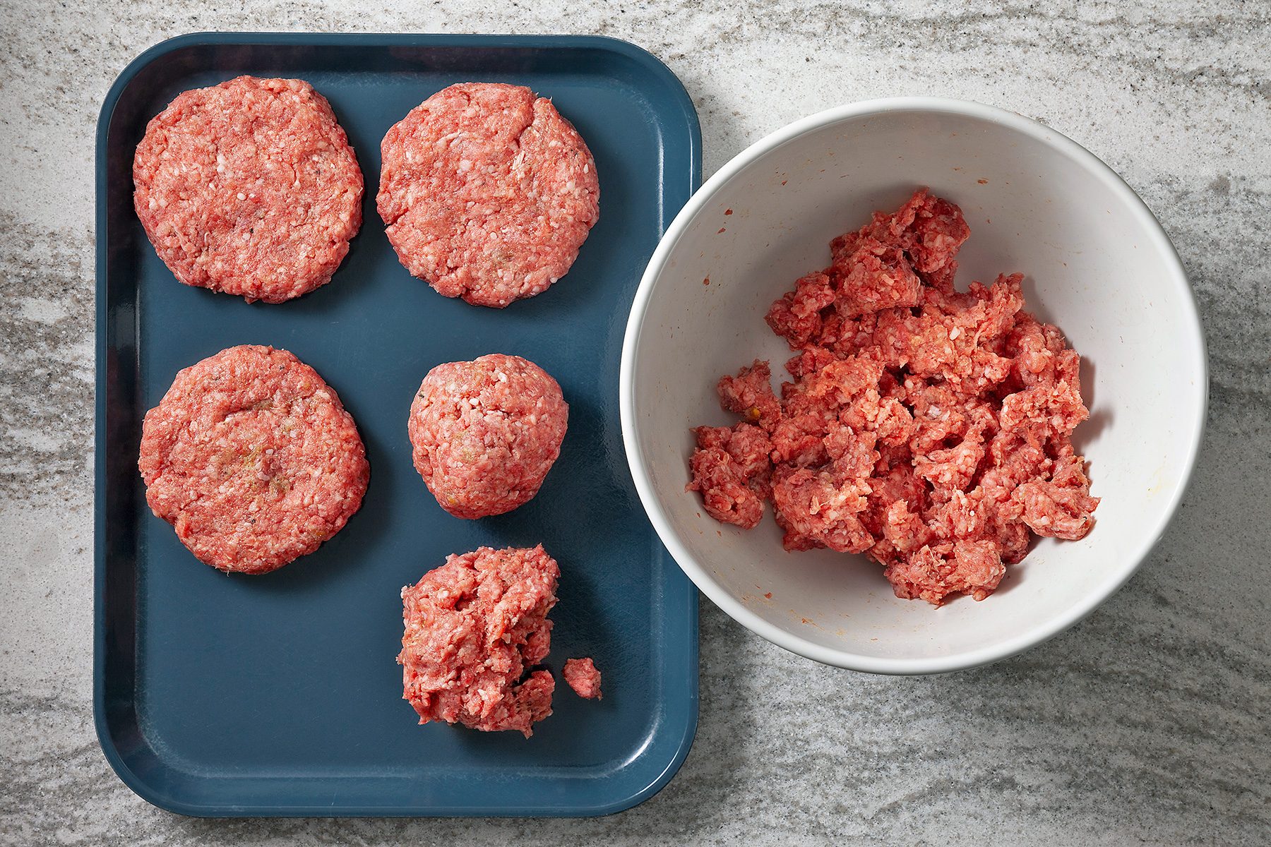 Ground meat is being prepared on a kitchen counter. Five formed patties are arranged on a blue tray to the left, while a white bowl on the right contains more raw meat ready to be shaped. The surface is a light gray textured countertop.