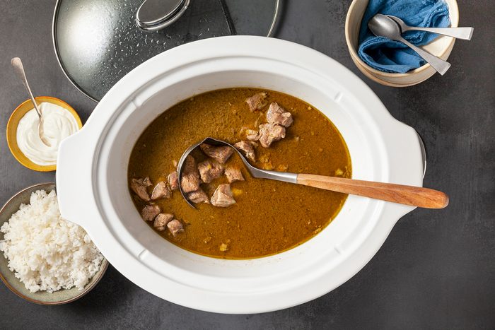 A white slow cooker filled with curry and chunks of meat, with a wooden ladle resting inside. Surrounding it are bowls containing steamed white rice, sour cream, a spoon, and a napkin. A lid is partially shown on the side.