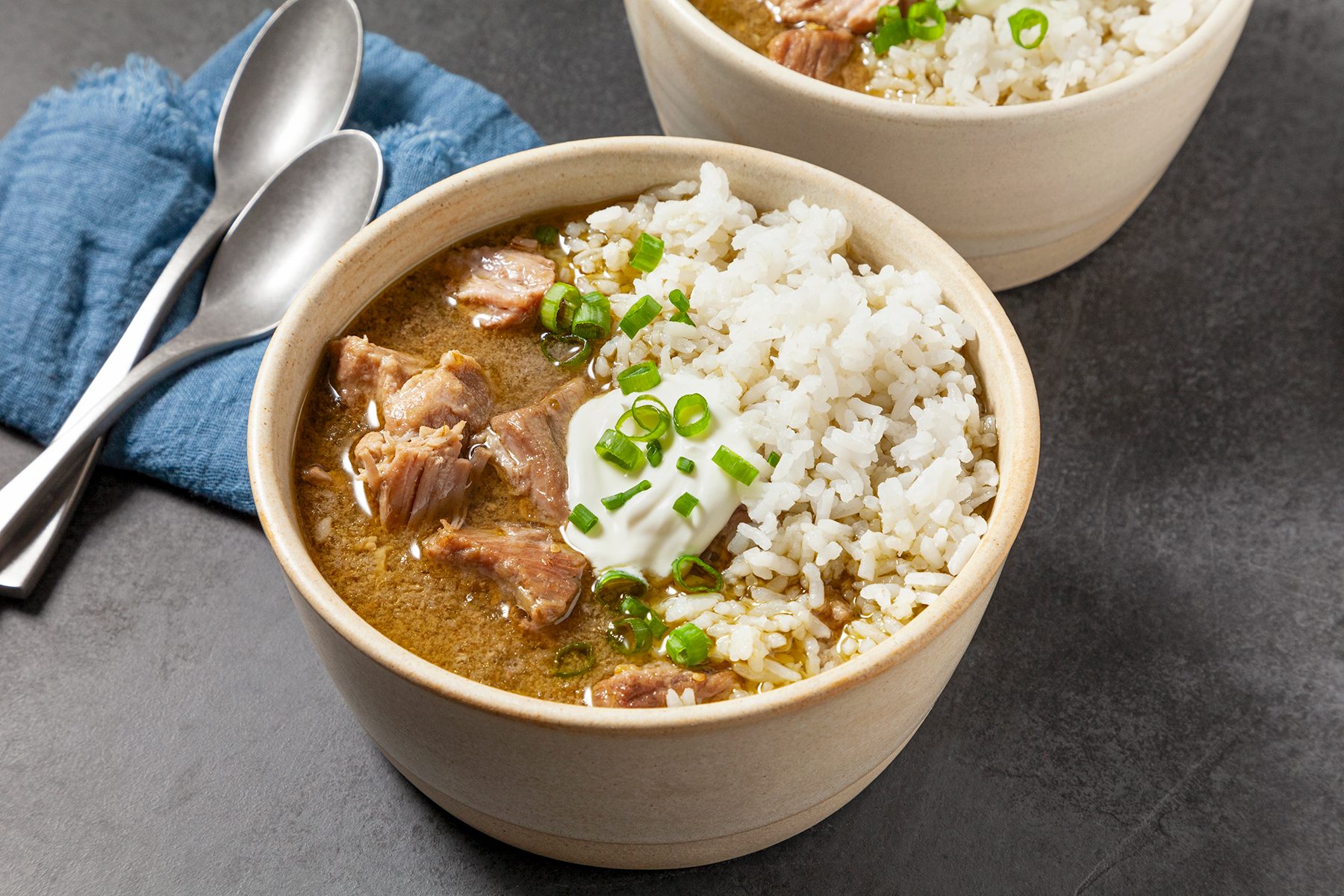 A bowl of rice and curry featuring chunks of meat, garnished with scallions and a dollop of sour cream. Two metal spoons rest on a blue cloth nearby, with another similar bowl partially visible in the background.