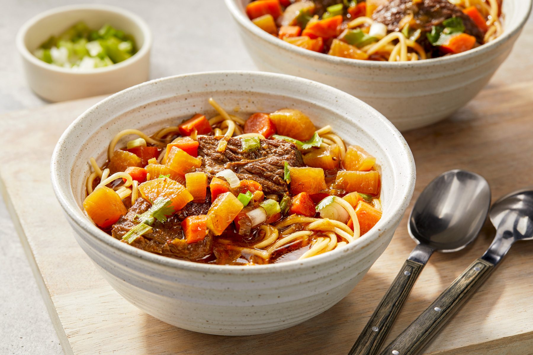 Horizontal AP; Pull shot of A bowl of beef noodle soup with carrots and potatoes sits on a wooden cutting board, along with two spoons with wooden handles; behind it is a second bowl of soup, partially visible; a small bowl of chopped green onions sits to the left of the frame.