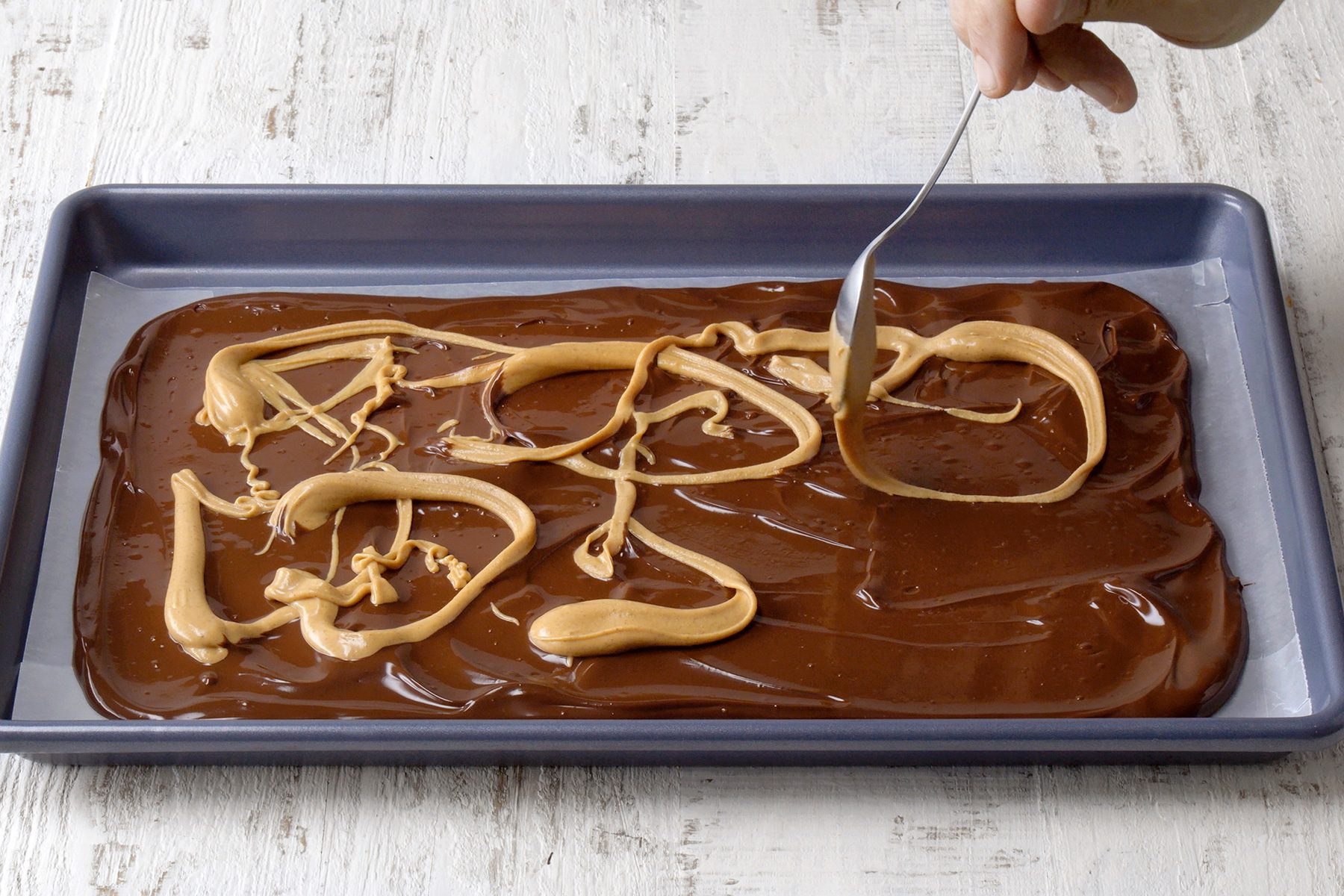 A baking tray holds smooth chocolate spread, with swirls of peanut butter on top. A hand uses a metal fork to further swirl the peanut butter into the chocolate, creating a marbled effect. The background is a textured white surface.