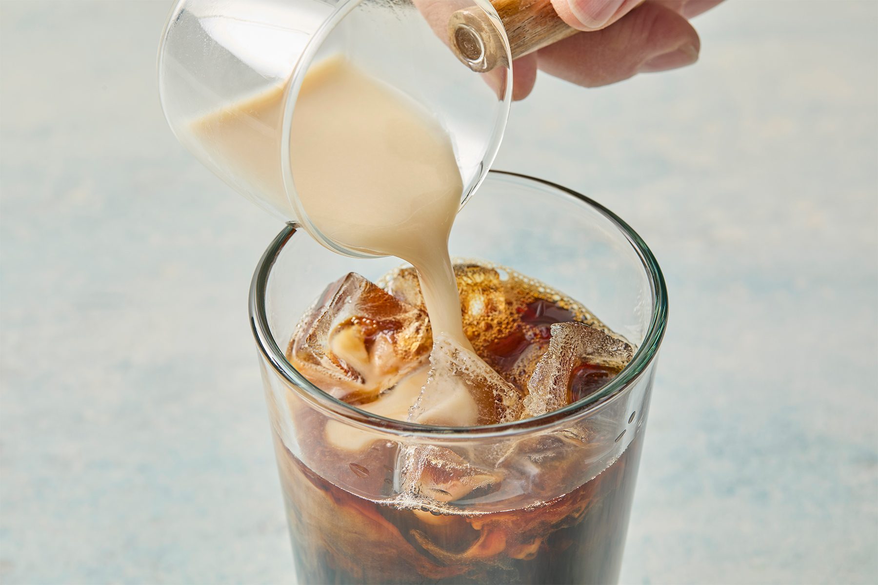 A close-up of a glass filled with iced coffee, into which a creamy liquid is being poured from a small glass pitcher. The background is a soft, light blue.
