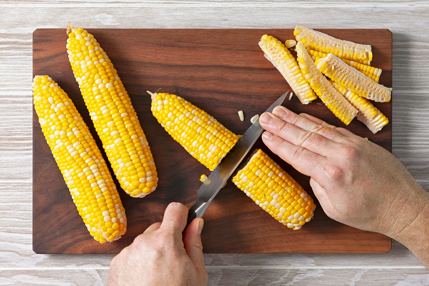 Overhead shot of remove to cutting board; cool; cut each corn cob in half widthwise; knife; chopping board; wooden surface;