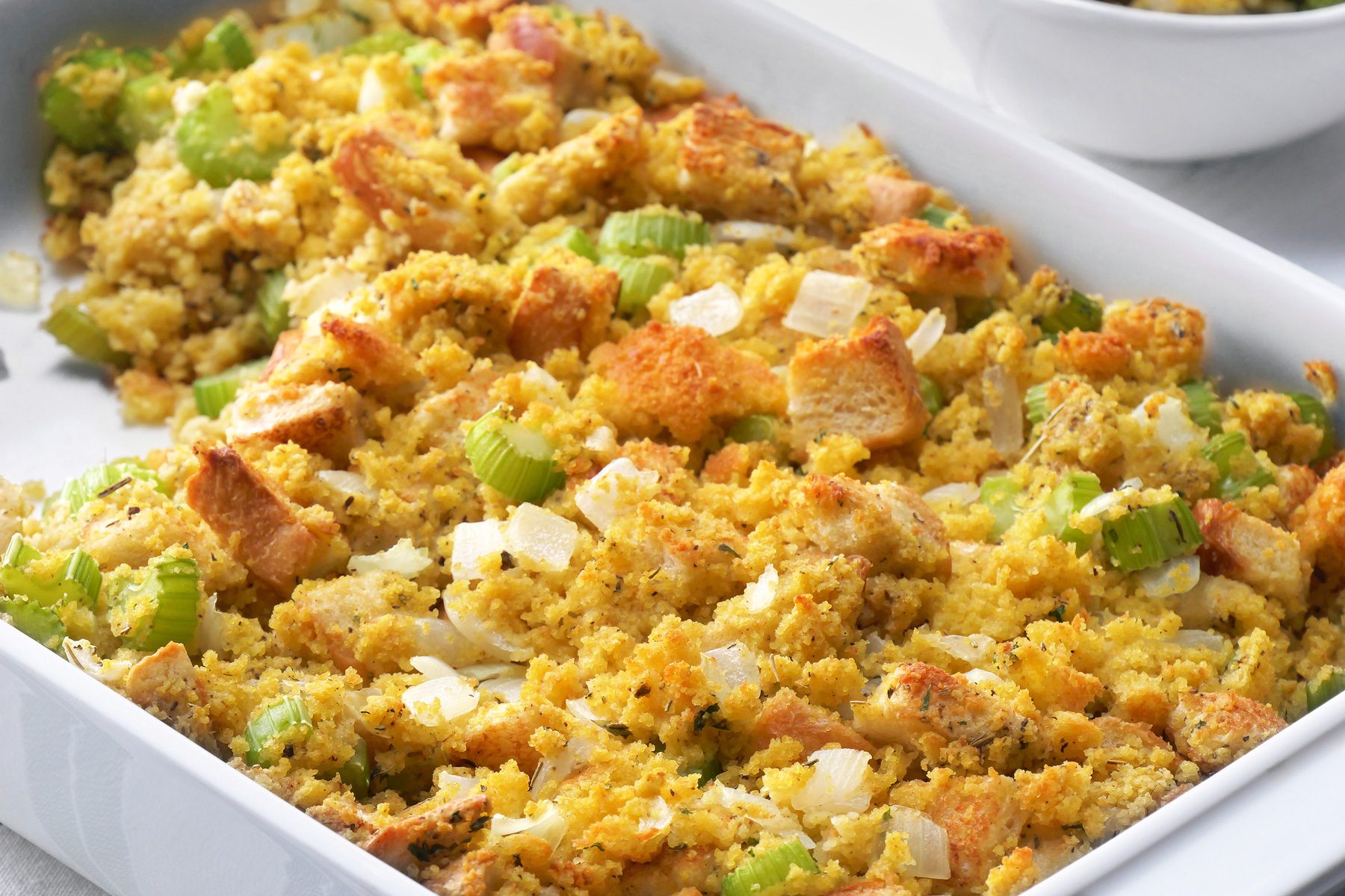 3/4th shot of a white baking dish filled with Cornbread Dressing is showcased, surrounded by two smaller bowls containing individual servings, The stuffing is packed with texture and flavor, featuring cubes of cornbread, chopped celery, and grated Parmesan cheese;