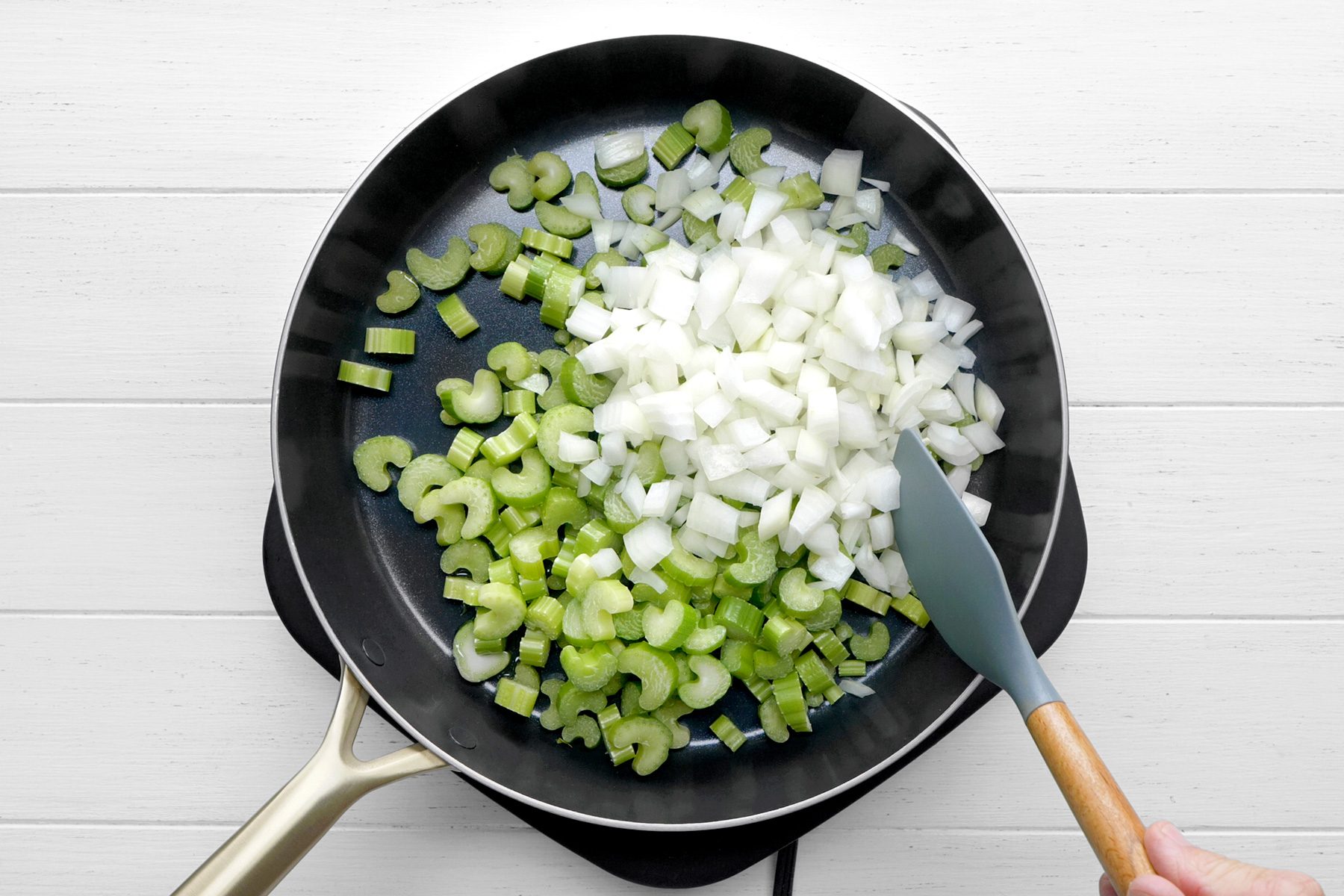 overhead shot of a black skillet on an electric stovetop, The skillet contains chopped celery and chopped onions, The celery is green and the onions are white, A spatula is being used to stir the vegetables, the vegetables are being sautéed;