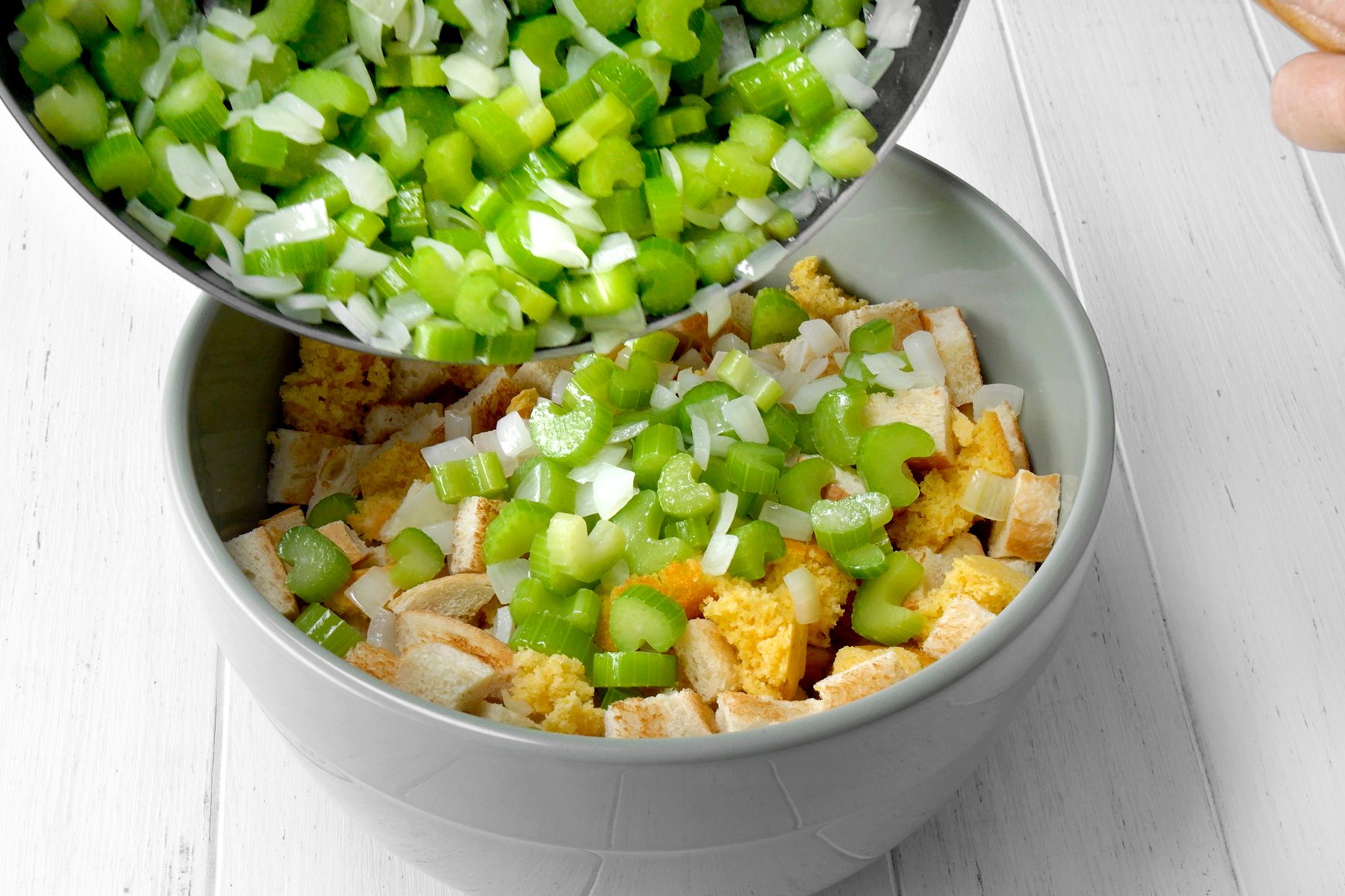 3/4th shot of a gray bowl filled with crumbled bread is shown, A person's hand is pouring chopped celery and onion into the bowl, The celery is green and the onion is white, these ingredients are being combined;