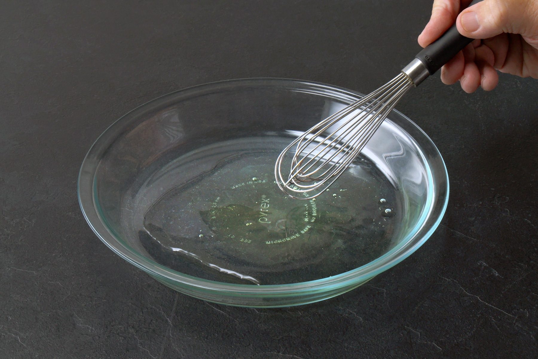3/4th shot of a hand holding a whisk and whisking a egg white mixture in a glass bowl, The liquid appears to be a light yellow color and has bubbles forming on the surface, The bowl is placed on a dark gray surface