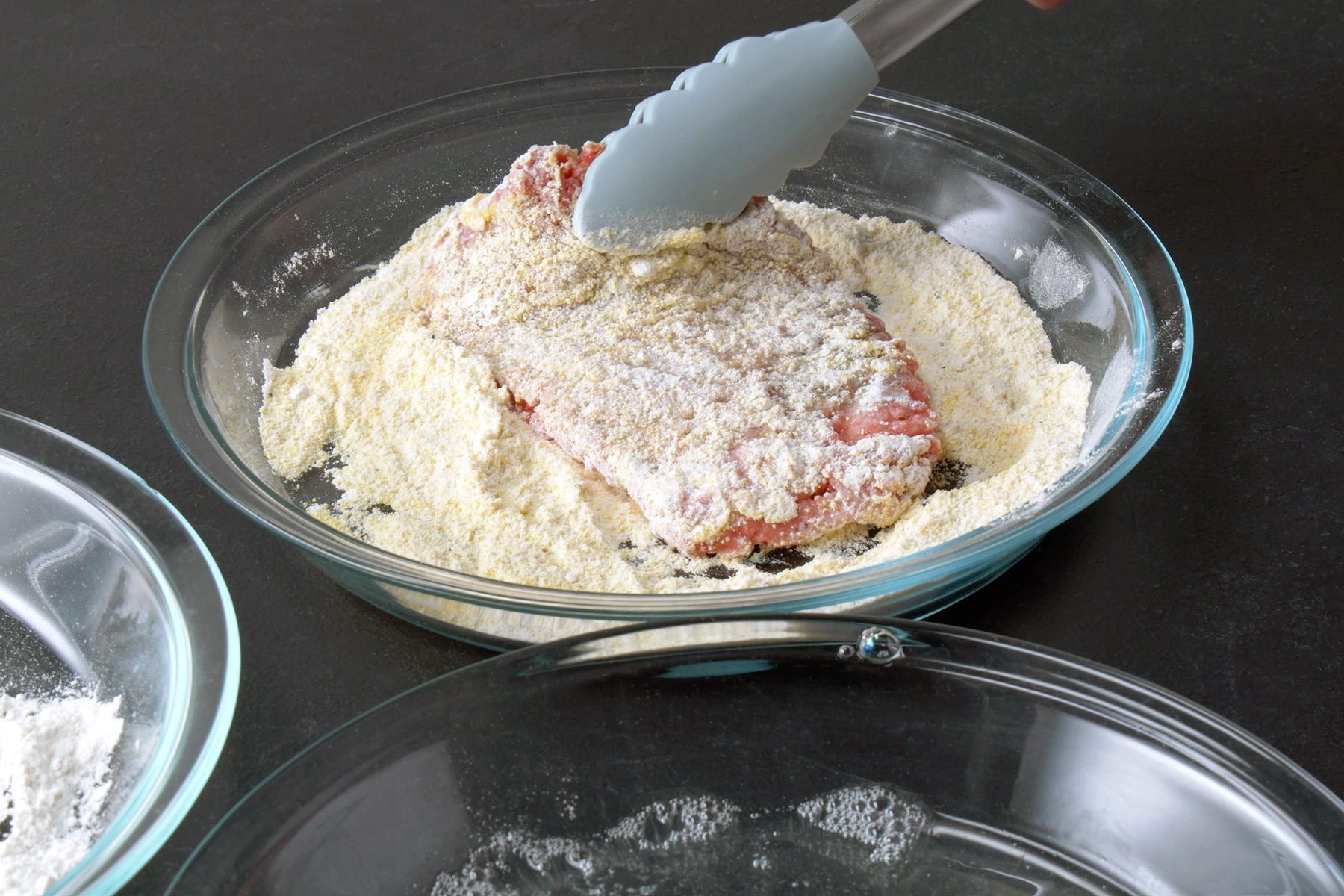 3/4th shot of a breaded meat patty being coated in a mixture of breadcrumbs and spices, A pair of tongs is being used to press the meat patty into the mixture, ensuring it is evenly coated, The meat patty is a reddish-brown color, and the breading mixture is a light brown color