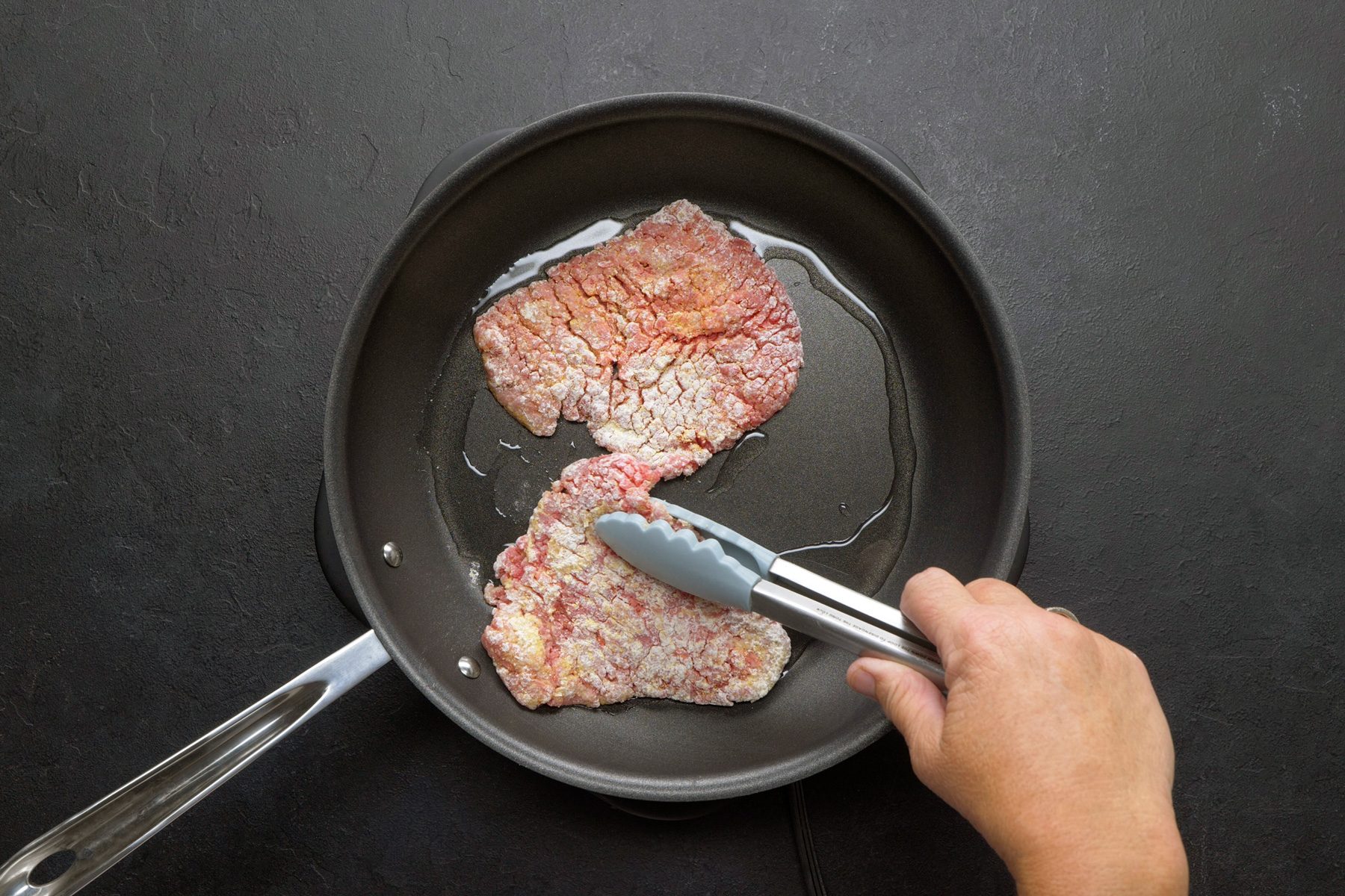 overhead shot of a breaded meat cutlet, likely a pork or beef steak, is being cooked in a skillet, The cutlet is coated in a golden brown breadcrumb mixture and is sizzling in hot oil, A pair of tongs is being used to flip the cutlet