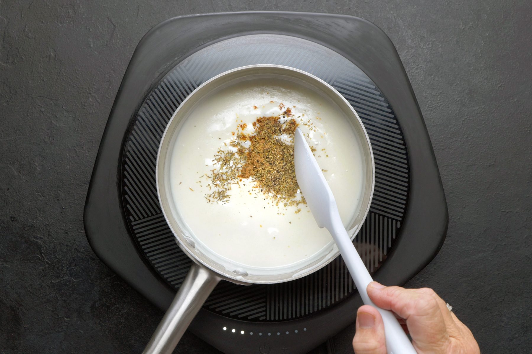 overhead shot of a saucepan on an electric induction cooktop, The saucepan contains a creamy white sauce, and a variety of dried herbs and spices are being stirred into it using a spatula, The herbs and spices include dried oregano, dried thyme, and possibly other spices, The sauce is simmering gently, and steam is rising from the saucepan