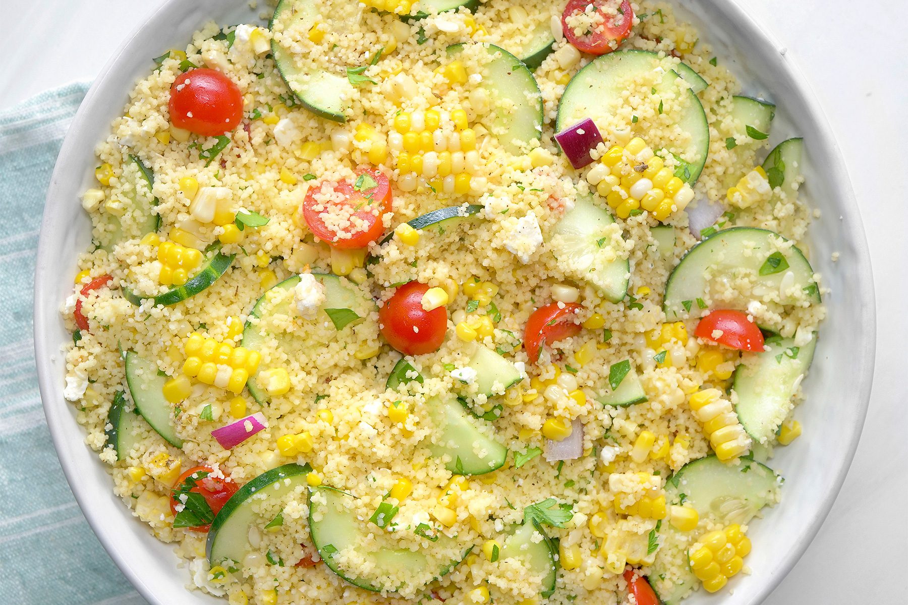 overhead shot of a white bowl is filled with a colorful couscous salad, The salad contains couscous, diced cucumbers, cherry tomatoes, crumbled feta cheese, and chopped fresh parsley, The ingredients are mixed together with a light dressing;
