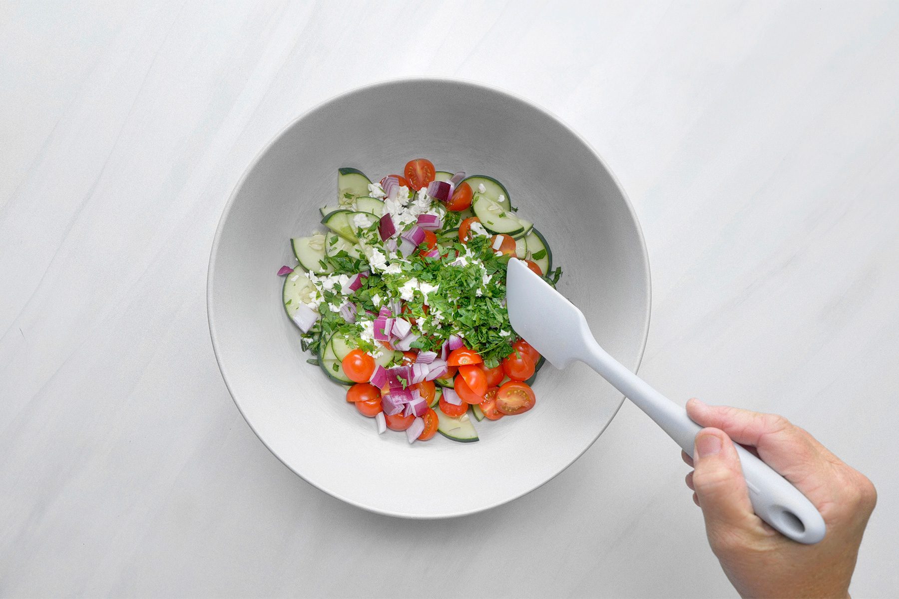 overhead shot of a white bowl contains a colorful salad of chopped cucumbers, cherry tomatoes, red onion, and crumbled feta cheese, Fresh parsley is being added to the salad, and a spatula is being used to gently toss the ingredients together;