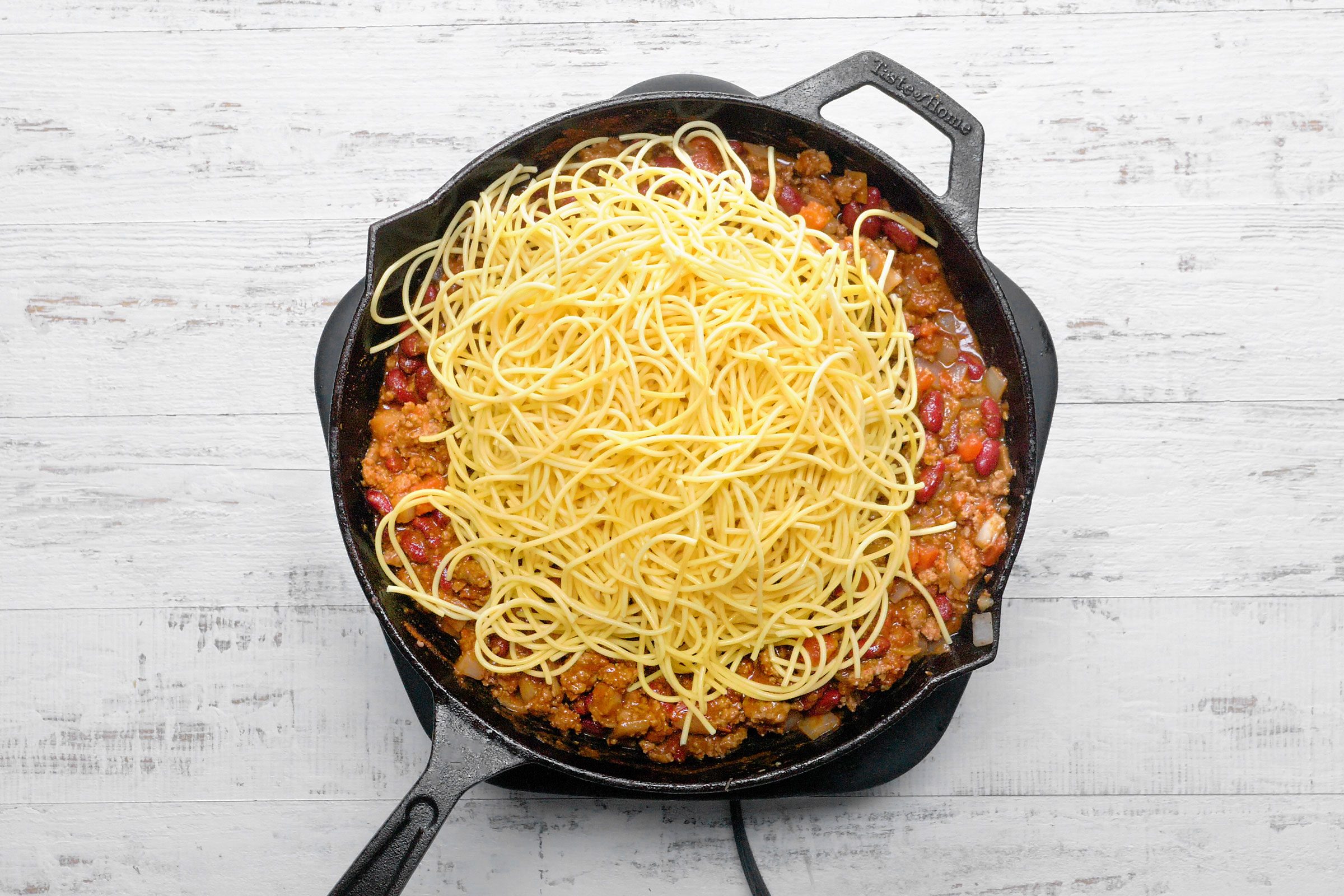 Drained spaghetti put into a skillet filled with bacon, beans, sauces and spices