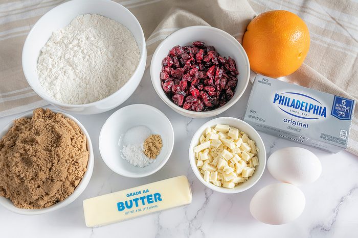 Baking ingredients on a marble surface: flour, dried cranberries, an orange, brown sugar, baking soda, eggs, white chocolate chips, a stick of butter, and a block of cream cheese.