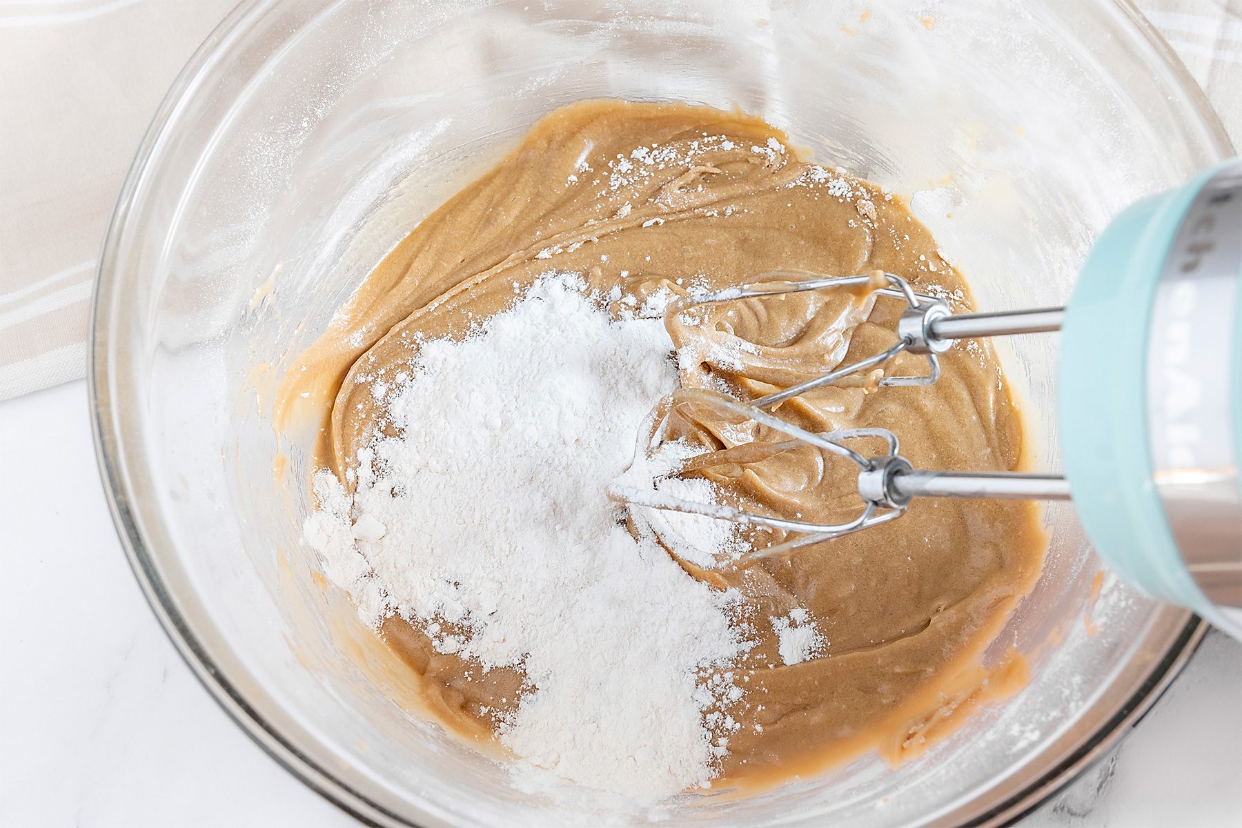 A glass bowl containing a mixture of creamy peanut butter is being mixed with an electric beater. A pile of white flour rests on top of the mixture, ready to be blended in. The background is a light-colored countertop.