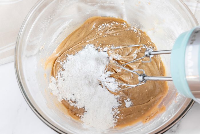 A glass bowl containing a mixture of creamy peanut butter is being mixed with an electric beater. A pile of white flour rests on top of the mixture, ready to be blended in. The background is a light-colored countertop.