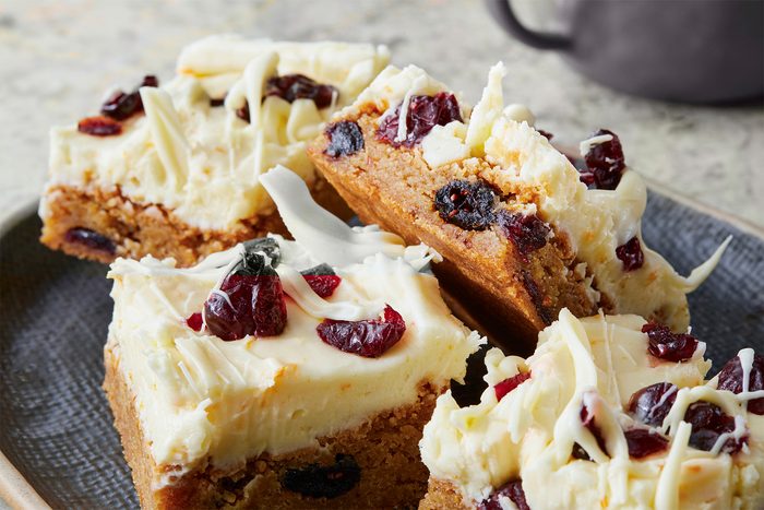 Four frosted blondies with dried cranberries and white chocolate shavings on top, served on a gray plate.