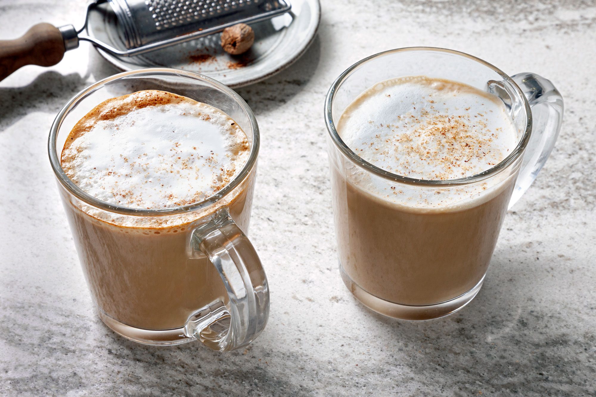 Two glass mugs filled with coffee and a sprinkle of nutmeg on top of the foam sit on a white countertop