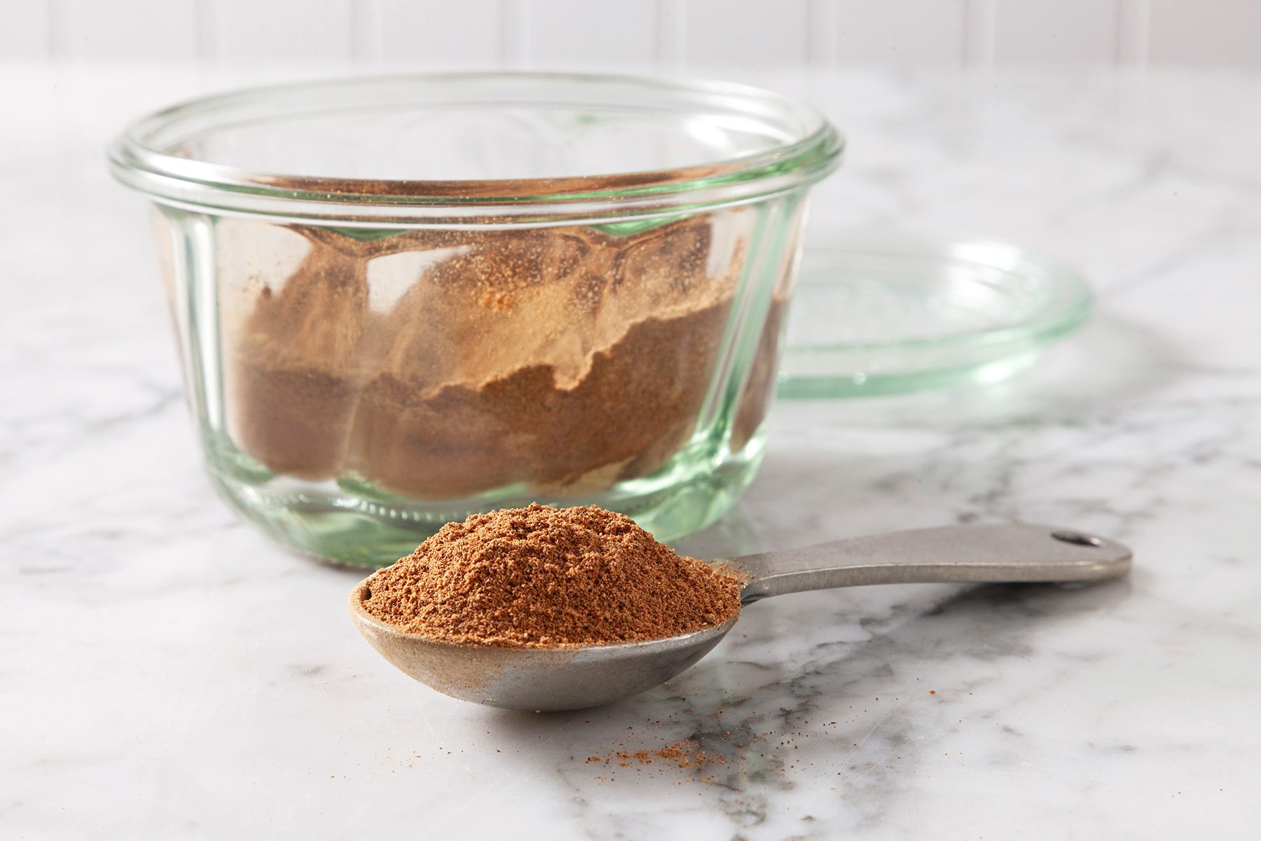 A glass jar filled with ground cinnamon sits on a marble countertop, with a metal spoon in front containing a heap of the same cinnamon powder. The jar lid is placed behind the jar.