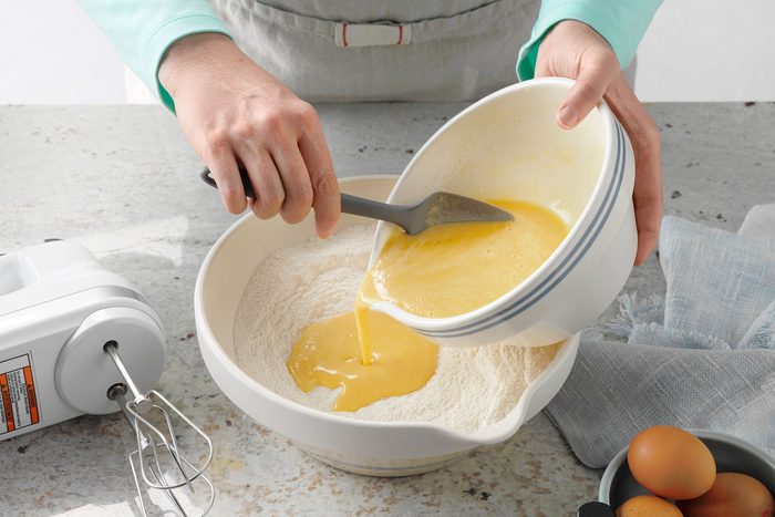 A person is pouring a bowl of beaten eggs into a larger bowl containing dry ingredients on a kitchen countertop. An electric mixer and some eggs are nearby.