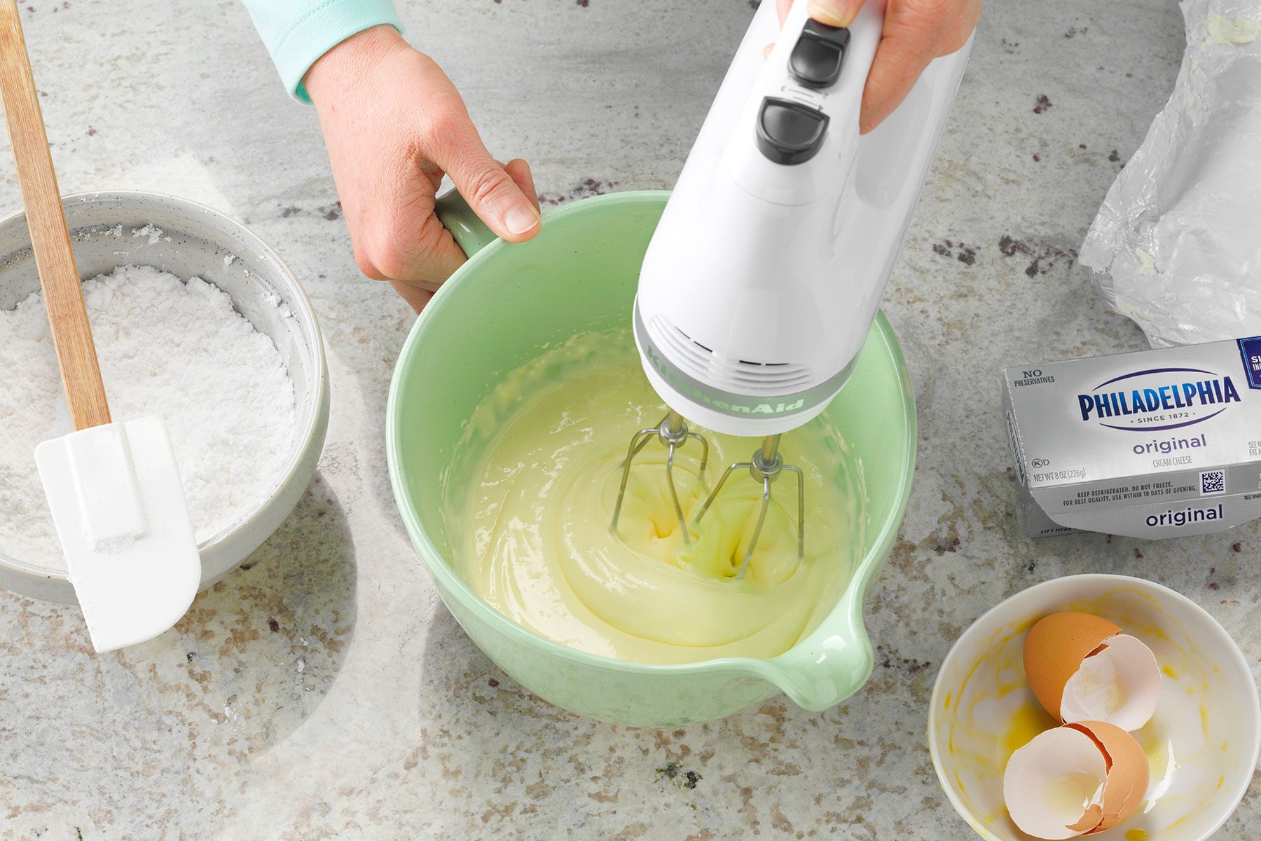A person uses a white electric hand mixer to blend ingredients in a light green bowl. Nearby are a rubber spatula, a bowl of flour, a package of cream cheese, and cracked eggshells on a countertop.