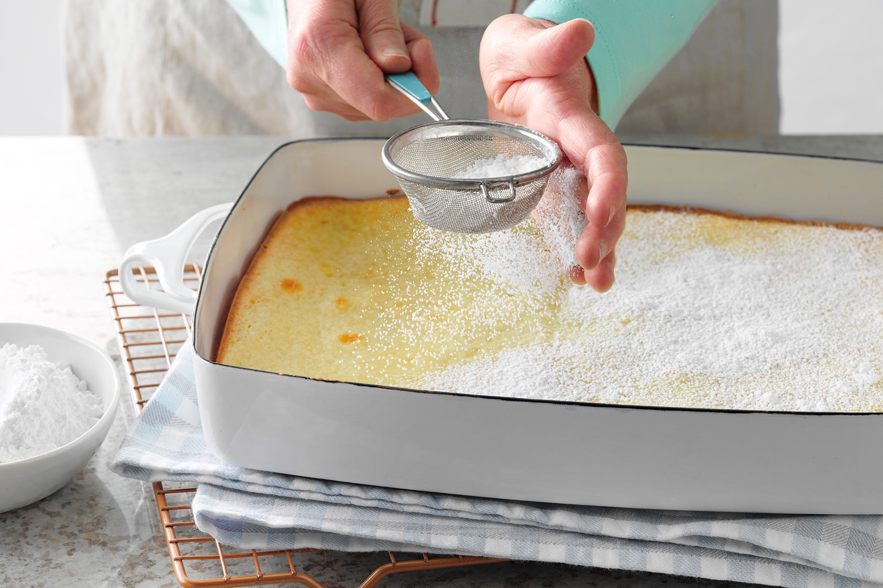 A person sprinkles powdered sugar over a freshly baked dessert in a white rectangular dish. The dish is resting on a checkered cloth and a cooling rack. A bowl of powdered sugar is nearby on the left.