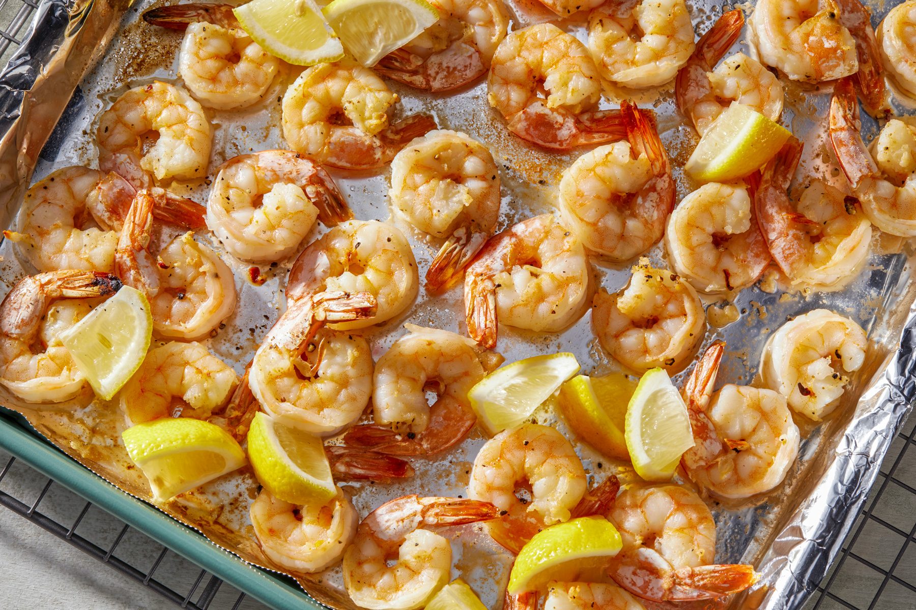 Horizontal AP pull shot of a baking sheet filled with cooked shrimp in a pale pink color, scattered across the sheet, with slices of lemon placed among them.