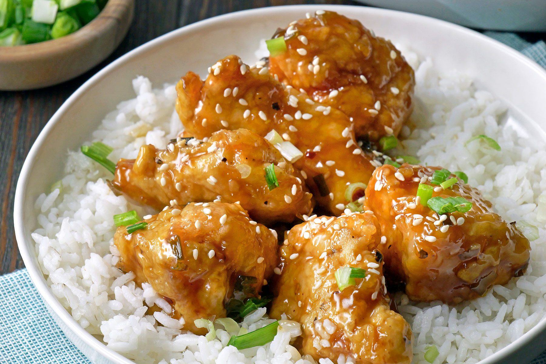 A bowl of white rice topped with glazed, golden-brown chicken pieces, garnished with sesame seeds and chopped green onions.