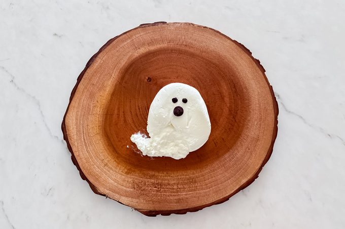 A dessert resembling a small ghost, made of a dollop of white cream with two black eyes and a black mouth, sits on a round wooden plate. The plate is placed on a white marble surface.